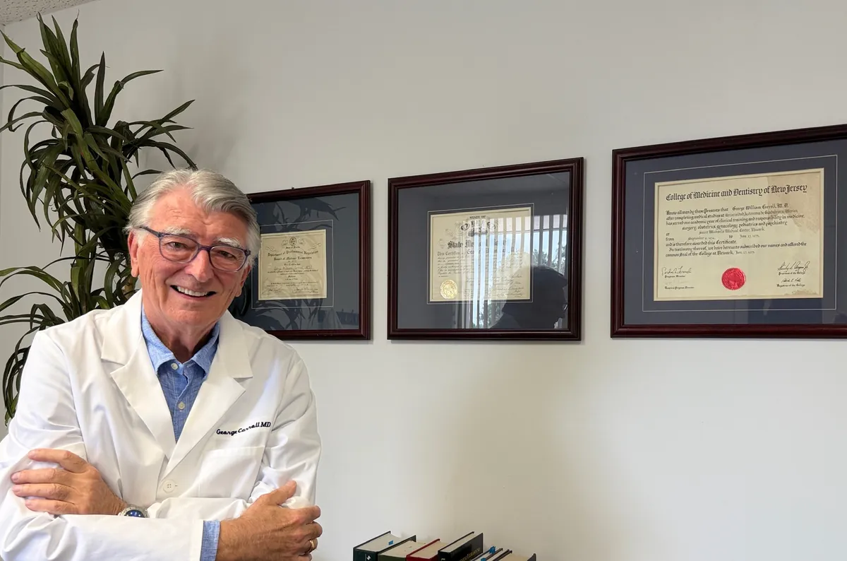 Dr. Carroll, arms folded and smiling, in front of his framed certificates